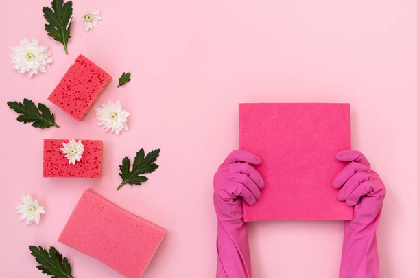 Mujer sosteniendo micro fibra toallita en las manos contra algunas esponjas de poliuretano, flores blancas y hojas verdes aisladas sobre fondo rosa pastel
 - Foto, Imagen