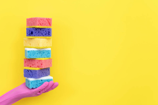 Mulher em luvas de borracha rosa segurando pilha de diferentes esponjas de prato de poliuretano para lavar na palma da mão isolada em fundo amarelo com espaço vazio para texto
 - Foto, Imagem