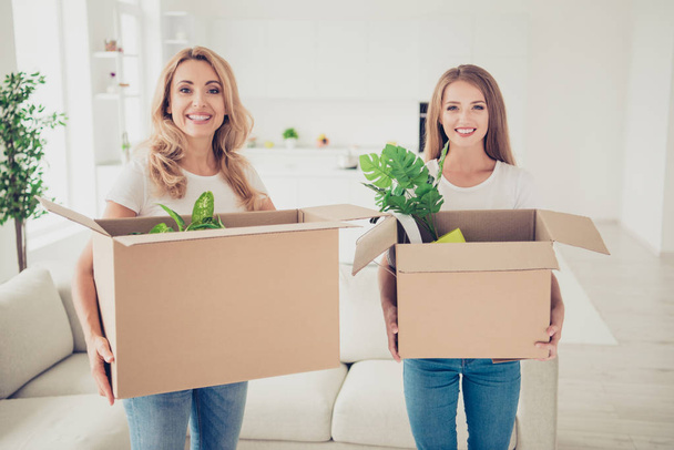 Close up photo two people mum and teen daughter with paper big boxes in arms ready to change decor do repair wear white t-shirts jeans in bright flat stand in front of comfortable sofa  - Foto, Bild
