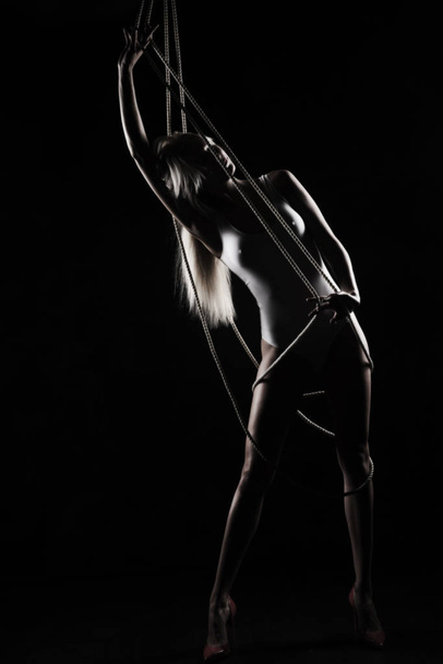 Beautiful slender blonde girl, wearing a white bodisuit and red stilettos, sensually plays and poses with the ropes. Black background. Artistic noir silhouette photo. Copy space. - Photo, image