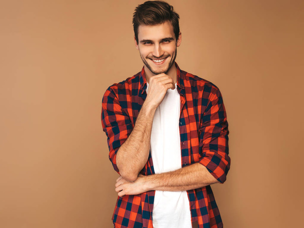 Portrait of handsome smiling stylish hipster lumbersexual businessman model dressed in red checkered shirt. Fashion man posing on golden background. Touching his chin - Valokuva, kuva