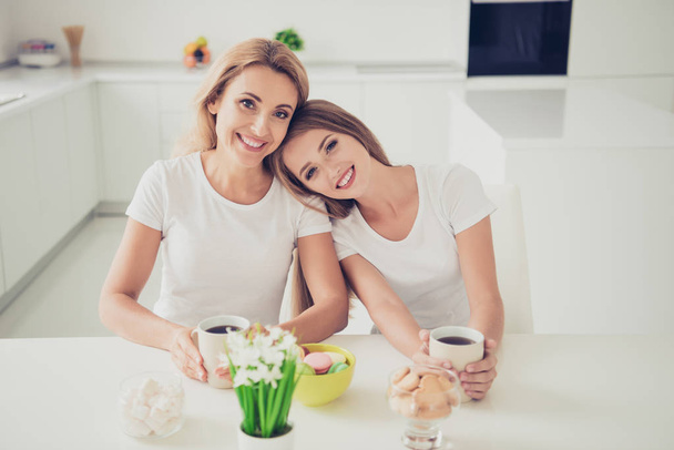 Close up photo two people mum and teenager daughter did house work duties drink tea coffee family moment head lovely lean on mommy shoulder wear white t-shirts jeans in bright flat kitchen - Foto, Bild