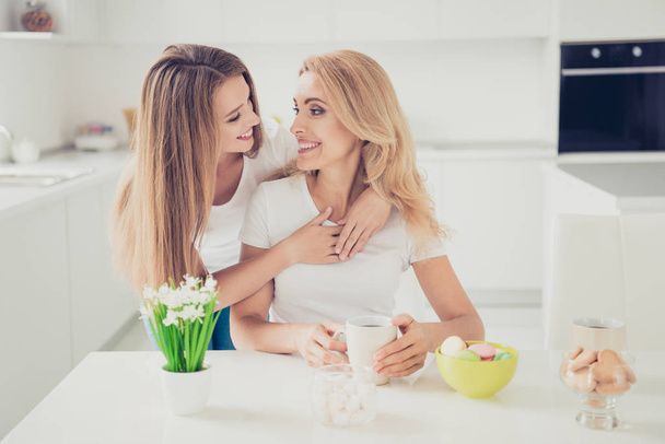 Close up photo piggy back two people mum and teenager daughter drink tea coffee family moment lovely lean on mommy look eyes woman holiday wear white t-shirts jeans in bright flat kitchen - 写真・画像