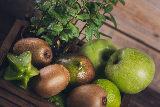 Fondo alimentario saludable. Concepto de comida saludable. Frutas verdes frescas en caja sobre fondo de madera oscura. Conjunto de frutas verdes para una dieta saludable y desintoxicación: manzana, lima, kiwi, mango, carambola y menta
. - Foto, Imagen
