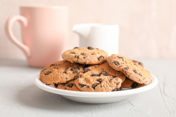 Bord met lekkere chocolade chip cookies en wazig kopje melk op grijze achtergrond, close-up - Foto, afbeelding