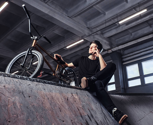 Bmx rider relaxing after practicing tricks with his bike in a skatepark indoors - Photo, Image