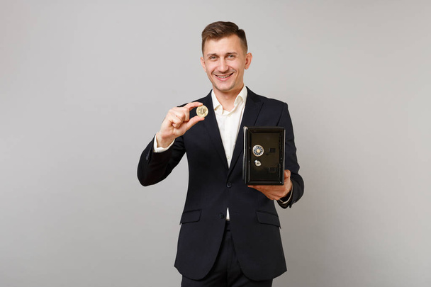 Smiling young business man in suit holding bitcoin, future currency, metal bank safe for money accumulation isolated on grey background. Achievement career wealth business concept. Mock up copy space - Photo, image