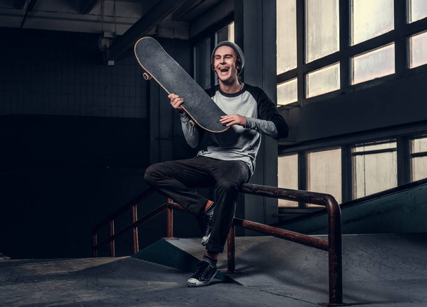 Cheerful man wearing a shirt and hat holding his board, sitting on a grind rail in skatepark indoors. - Photo, Image