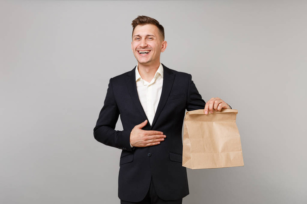 Laughing business man hold brown clear empty blank craft paper bag for takeaway mock up isolated on grey background. Achievement career wealth business concept. Food products delivery to work office - Photo, Image