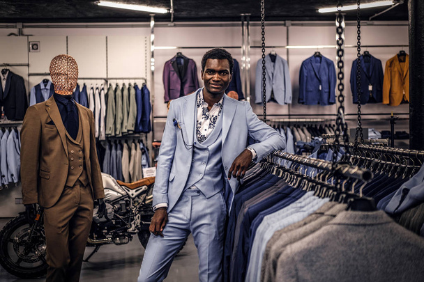 Homem afro-americano elegantemente vestido apoiando-se em um manequim em uma loja de moda masculina clássica
. - Foto, Imagem