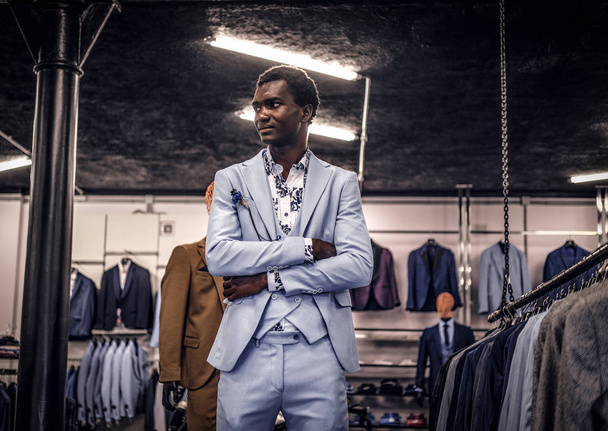 Homme afro-américain vêtu d'un costume élégant bleu posant avec les bras croisés dans un magasin classique de vêtements pour hommes
. - Photo, image