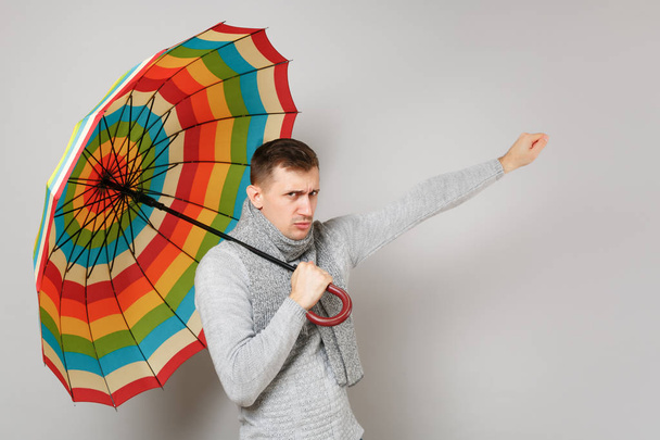 Upset young man in gray sweater, scarf holding colorful umbrella pointing hand isolated on grey background. Healthy fashion lifestyle, people sincere emotions, cold season concept. Mock up copy space - Photo, Image