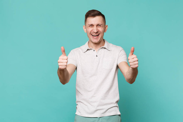 Portrait of laughing happy young man in casual clothes standing, showing thumbs up isolated on blue turquoise wall background in studio. People sincere emotions, lifestyle concept. Mock up copy space - Photo, Image