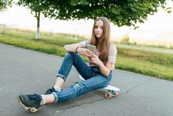 Girl teenager 9-11 years old, sits on board in hands of phone. In summer in city in casual jeans and a pink T-shirt. Communication in the Internet, in hands of holding a smartphone. Smiles and poses. - Фото, изображение