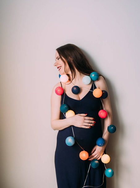 Pregnant woman in dark blue dress with christmas lanterns laughs - Photo, Image