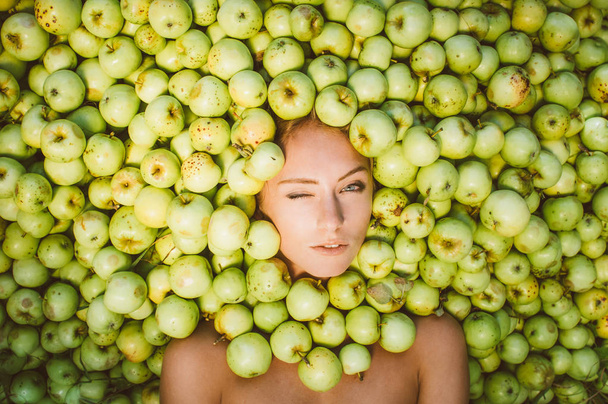 Porträt eines schönen Mädchens, das in den grünen Äpfeln liegt, Äpfel in der Nähe des Gesichts, ein geschlossenes Auge - Foto, Bild