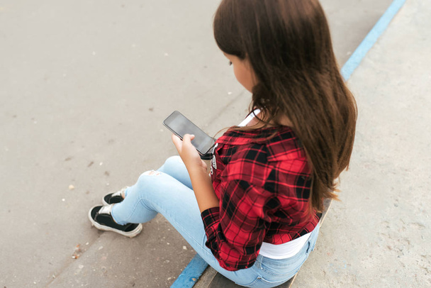 Girl teenager 11-12 years old sits on skateboard in park summer against background concrete stairs. In hands smartphone writes message in Internet application. Teenagers in social networks. Back view. - Φωτογραφία, εικόνα