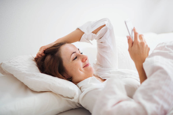 A young woman lying in bed indoors in the morning in a bedroom, using smartphone. - Valokuva, kuva