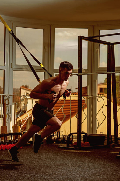 Man with nude torso, and muscular chest in gym enjoy training, trx. Sportsman, athlete, muscular macho does exercise with trx loops, window on background. Sport and gym concept. - Фото, изображение