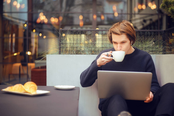 Man drinking cup of coffee in a restaurant on the terrace. Freelancer outdoor with a laptop, has breakfast with croissants. Businessman looks and dreams. Man with laptop . Wireless headphones.  - Fotoğraf, Görsel