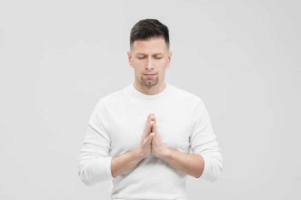 A man in white clothes on a white background with his hands folded on his chest. Meditation and prayer, yoga. - Foto, imagen
