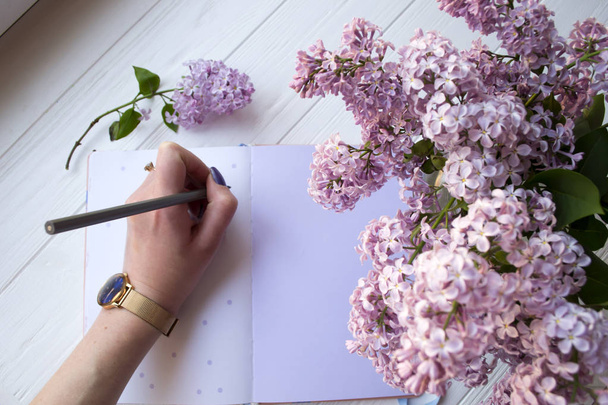 Lilac and notepad on a white desktop. Woman is writing in a notebook. Top view. - Foto, Bild