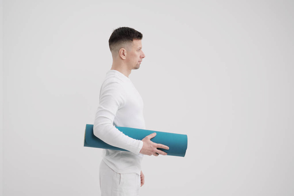 A male teacher and trainer in white clothes holding a yellow-blue yoga mat in his hands, preparing for a fitness class. - Foto, Imagen