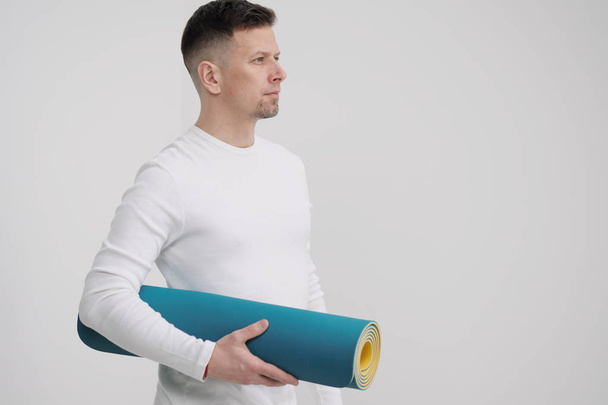 A male teacher and trainer in white clothes holding a yellow-blue yoga mat in his hands, preparing for a fitness class. - Φωτογραφία, εικόνα