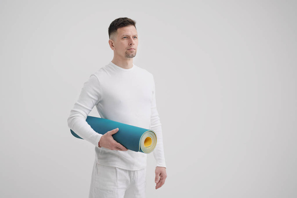 A male teacher and trainer in white clothes holding a yellow-blue yoga mat in his hands, preparing for a fitness class. - Fotoğraf, Görsel