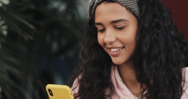 Happy african american girl sitting on the bed and chatting with her friends on her smartphone - Materiał filmowy, wideo