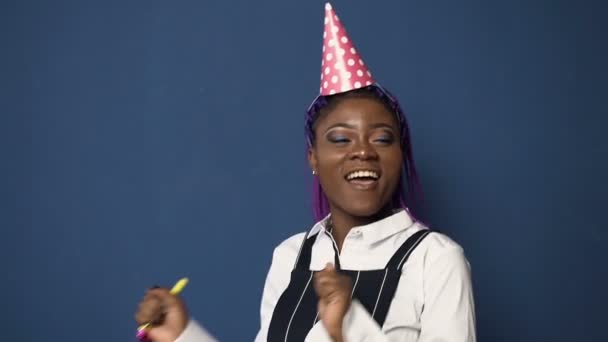 Happy african woman in pink birthday hat, white shirt dancing and whistling in birthday horn on the blue background. - Imágenes, Vídeo