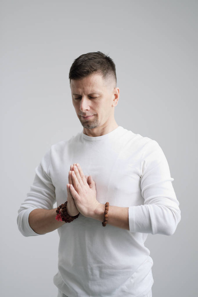 A man in white clothes on a white background with his hands folded on his chest. Meditation and prayer, yoga. - Zdjęcie, obraz