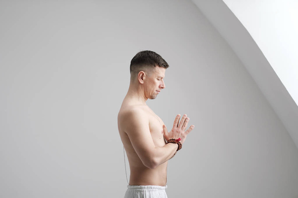 Un uomo vestito di bianco e a petto nudo su uno sfondo bianco con le mani piegate sul petto. Meditazione e preghiera, yoga
. - Foto, immagini