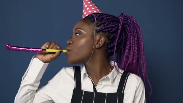 Side view of african young woman in hat dancing whistling in birthday horn on the blue background. - Filmmaterial, Video