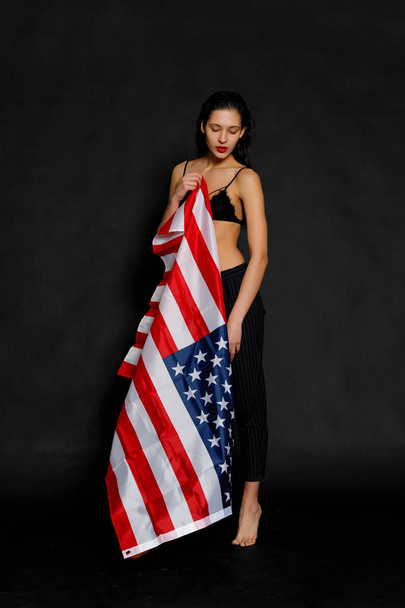 Portrait of proud female athlete wrapped in American Flag against black background. Muscular young woman looking confidently at camera. - Fotografie, Obrázek
