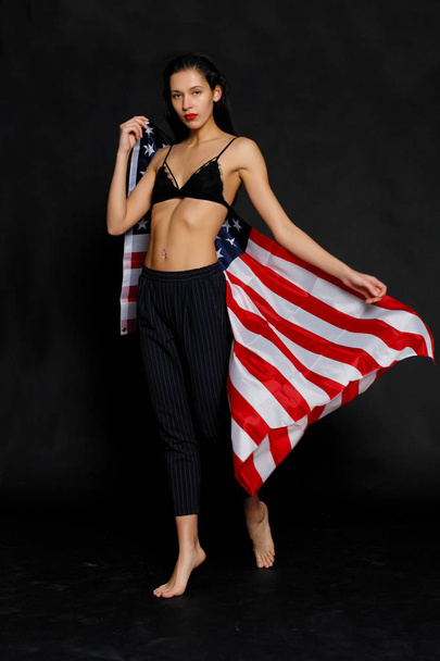 Portrait of proud female athlete wrapped in American Flag against black background. Muscular young woman looking confidently at camera. - Photo, image