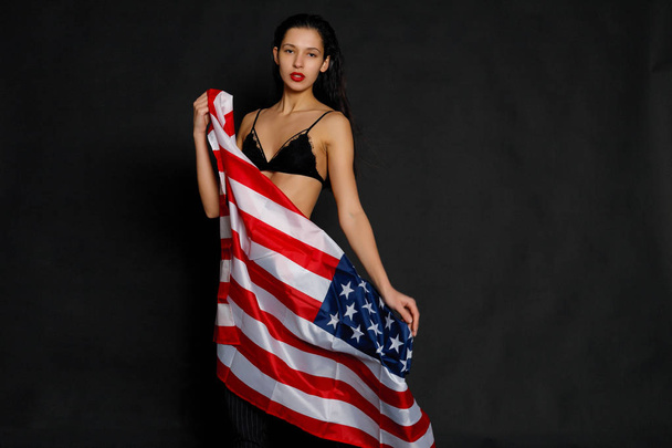 Portrait of proud female athlete wrapped in American Flag against black background. Muscular young woman looking confidently at camera. - Fotó, kép