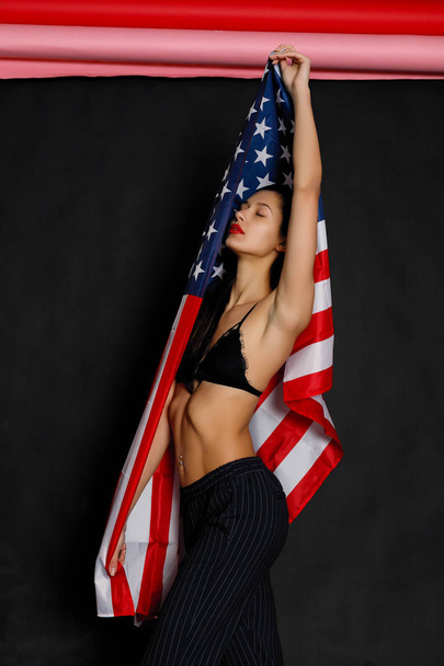 Portrait of proud female athlete wrapped in American Flag against black background. Muscular young woman looking confidently at camera. - Foto, Bild