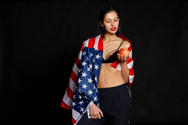 Portrait of proud female athlete wrapped in American Flag against black background. Muscular young woman looking confidently at camera. - Фото, изображение