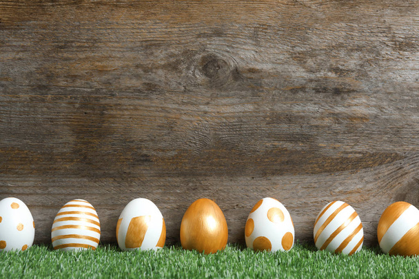 Line of traditional Easter eggs decorated with golden paint on green lawn near wooden wall, space for text - Fotó, kép