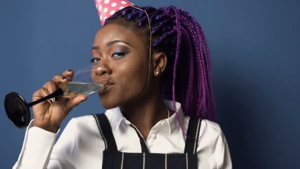 schöne afrikanische Mädchen mit Geburtstagshut trinken shampanage aus dem Glas auf dem blauen Hintergrund. - Filmmaterial, Video