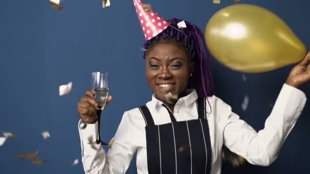 Happy young african woman in birthday hat with champanage glass in one hand and balloon in another dancing while confetti flying down on the blue background. - Filmati, video