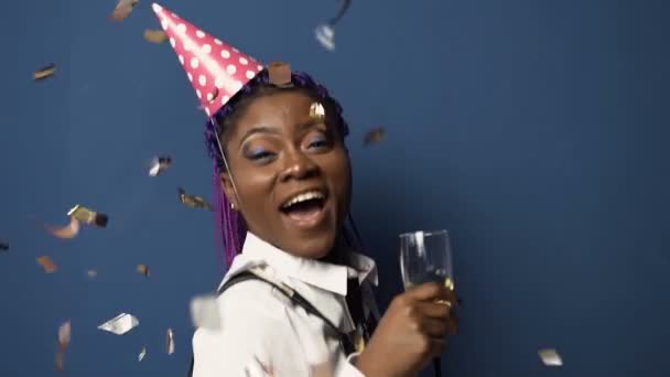 Attractive african girl in hat with champanage glass in hand, having fun and dancing while confetti flying down on the blue background. - Footage, Video