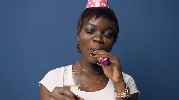 Attractive african woman in birthday hat holding champanage glass and whistling in birthday horn on the blue background. - Кадри, відео