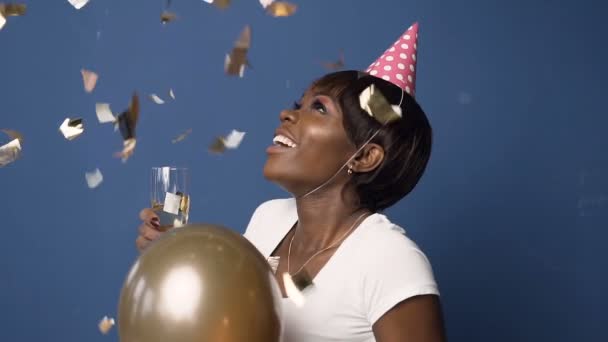 Happy african girl in hat with champanage glass in one hand and balloon in another, looking to the confetti which flying down on the blue background. - Footage, Video