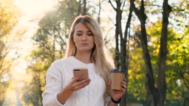 Stylish young cute smiling girl using modern smart phone while walking in the city park. Lady typing text message on cellphone and drinking coffee to go, outdoor, at sunset. - Filmati, video