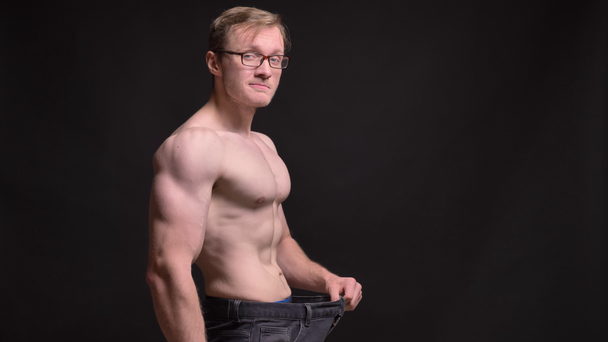 Ritratto di profilo di giovane uomo nudo muscoloso in occhiali che mostra felicemente i risultati della perdita di peso in macchina fotografica su sfondo nero
. - Filmati, video