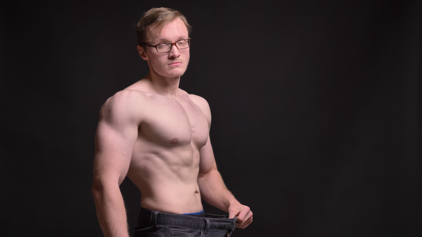 Portrait in profile of naked young muscular man in glasses gladly showing the results of losing weight into camera on black background. - Footage, Video