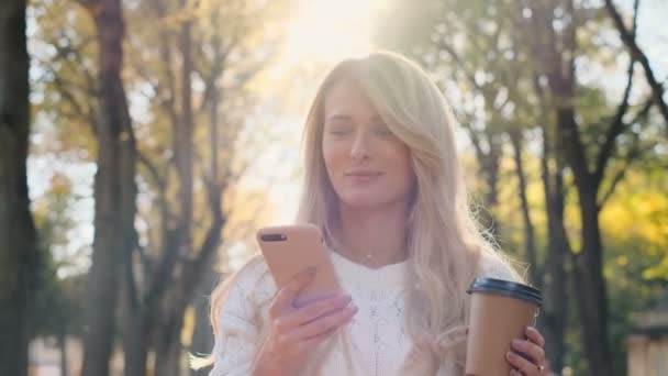 Beautiful young hipster girl wearing white sweater using modern smart phone while walking at break in the city park, woman employer typing text message on cellphone outside, portrait, slow motion - Video