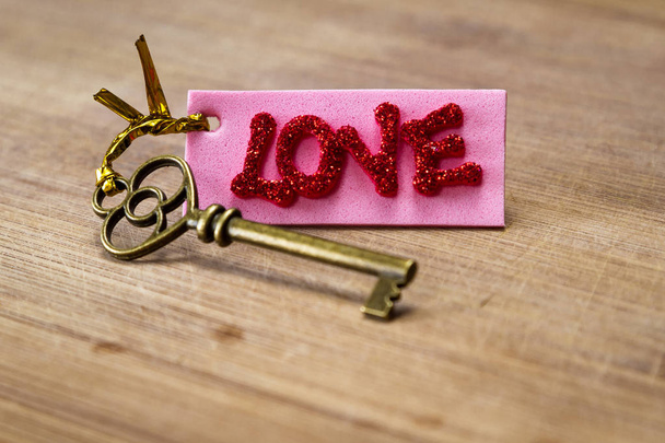 conceptual image using an old key with a pink tag with red glitter letters spelling the word love over a wooden background - Photo, Image